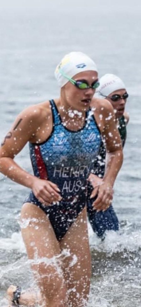 A swimmer sprinting out of the water during a competition, wearing a swim cap and swimsuit marked 'HENRY AUS' while splashing water around.